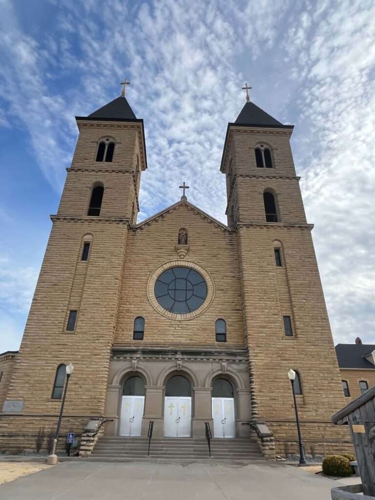 Image of St. Fidelis Catholic Cathedral in Victoria, KS. Image is from the street view looking upward at the twin spires that point to the sky, which overshadow the center portion of the building where a cross is centrally located. The front steps from the street rise up to three doors at the front.