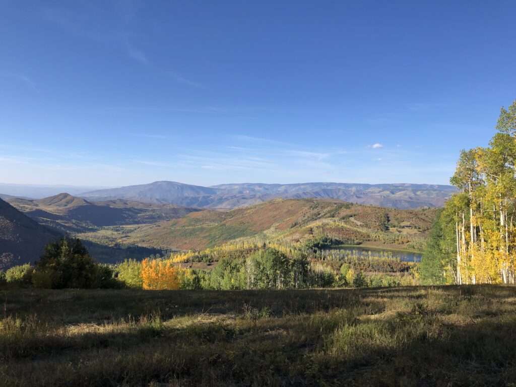 Sunrise from the top of a mountain looking down into the distance. There is a large meadow that goes downhill toward a small lake with mountains behind it. It is fall and the leaves are changing color