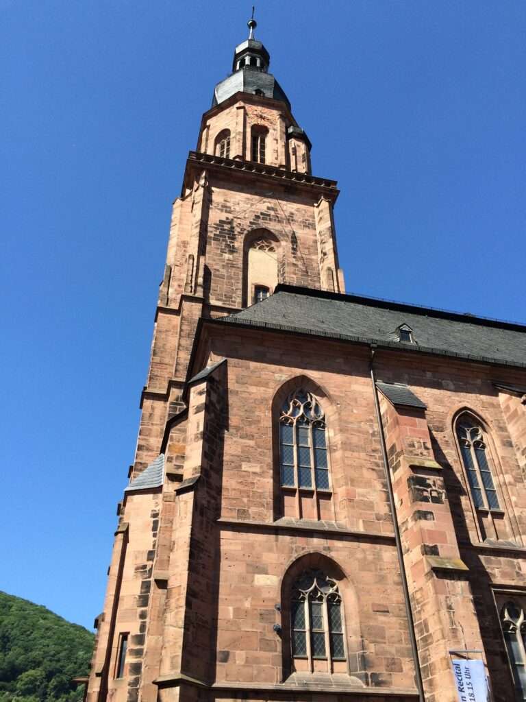 Picture of the exterior of the Church of the Holy Ghost in Heidelberg, Germany. It is a large stone church from the 1300's with a view of the steeple looking straight up.