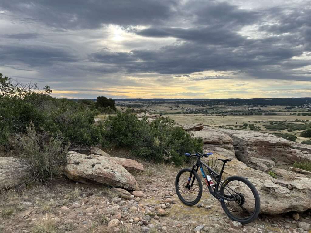 Sunrise looking down from the top of a butte with a cloudy sky. In the foreground is a mountain bike resting next to a rock and some scrub oak.
