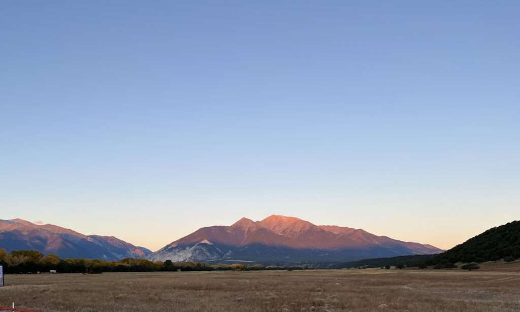 A picture of a large mountain in the vast distance with the rays of the sunrise shining on the face of the mountain. A long meadow stretches out to the base of the mountain.
