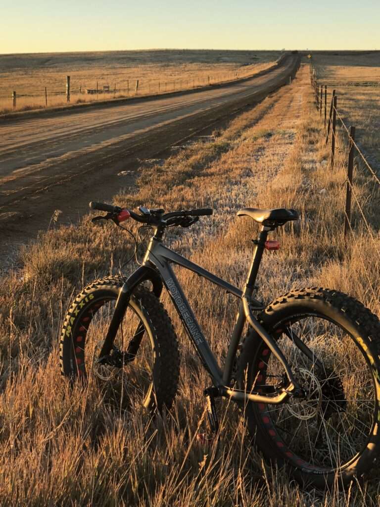 A picture of a fatbike leaning up agains a fence along a long dirt road that goes off into the distance. It is the moment of sunrise and the light is shining from the horizon