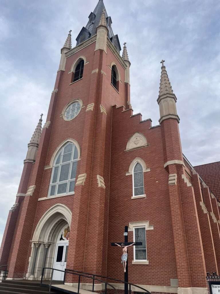 Picture of an old brick and stone church building with tall spires pointing upward. The picture is from the vantage point of being on the street looking upward as the building points to the sky.