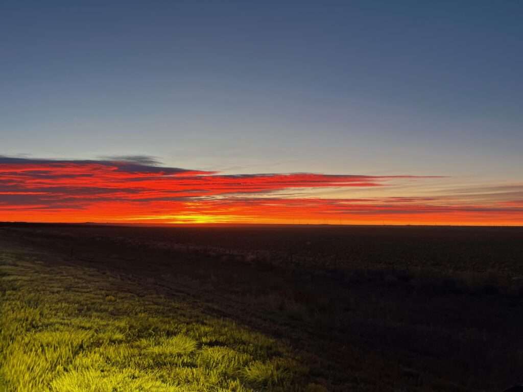 Picture of an early sunrise over Vona, CO. The picture has a long distance view of the horizon with the sun about to cross over. There are grassy plains as far as the eye can see and low colorful clouds on the horizon